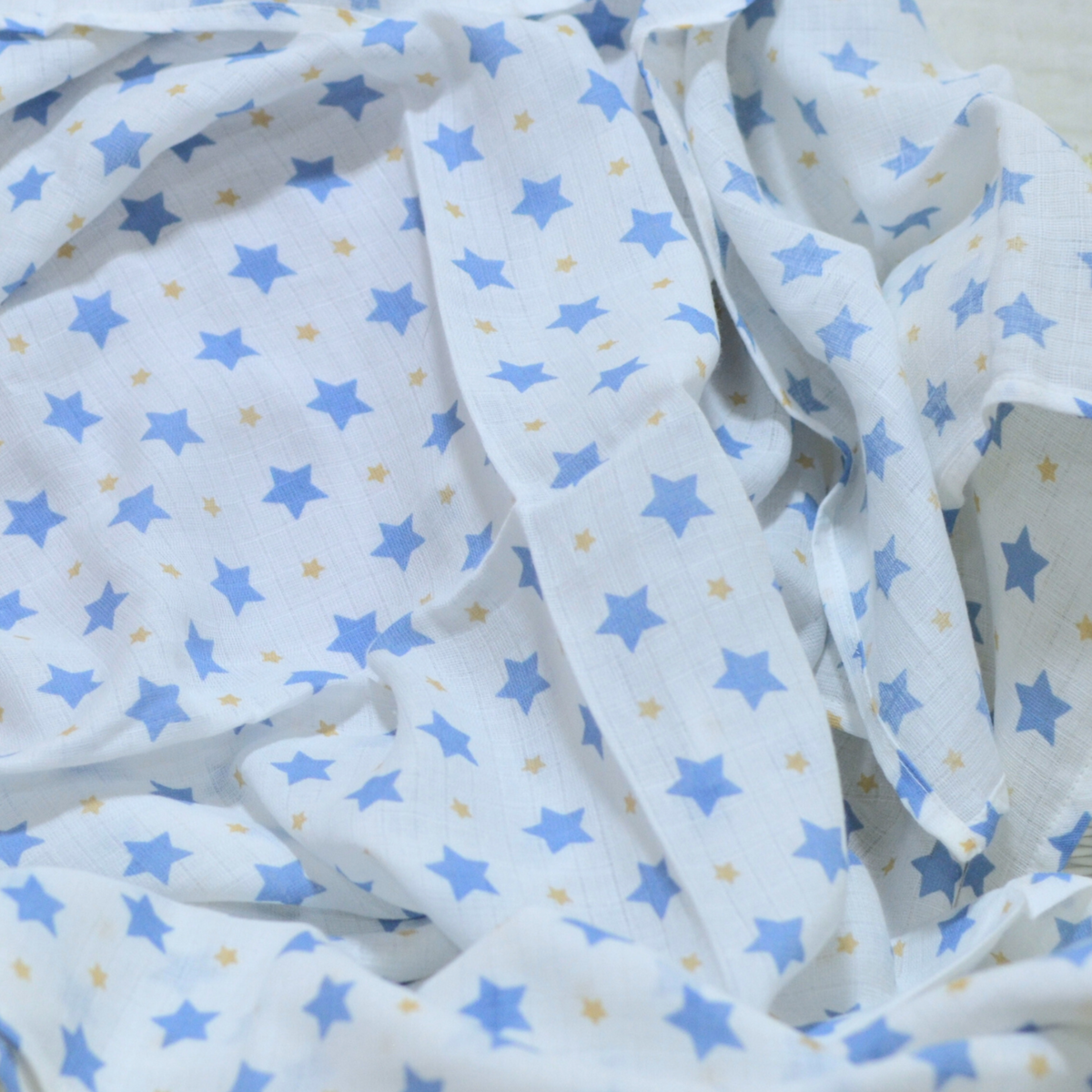 A white muslin baby blanket or swaddle with a pattern of blue stars and small yellow stars, draped in soft folds.